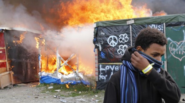 Un inmigrante se protege el rostro al pasar junto a chabolas en llamas. Foto: EFE